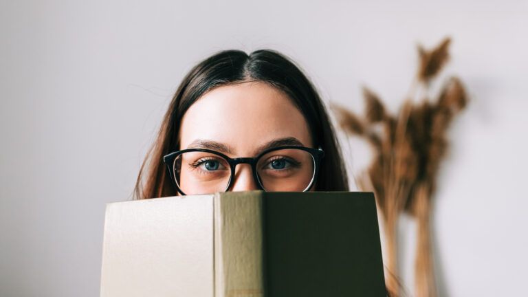 Woman with glasses looking over her book cover while doing her reading habit