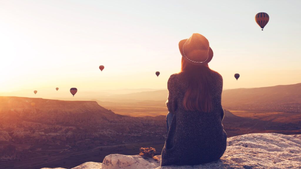 A woman sits on a hill, watching hot-air balloons