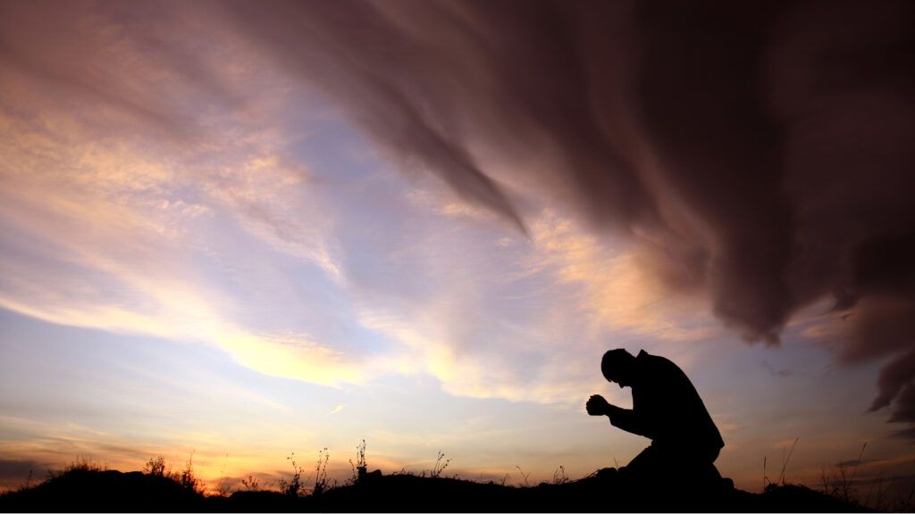 A silhouette of a man praying outdoors against a colorful sunset.