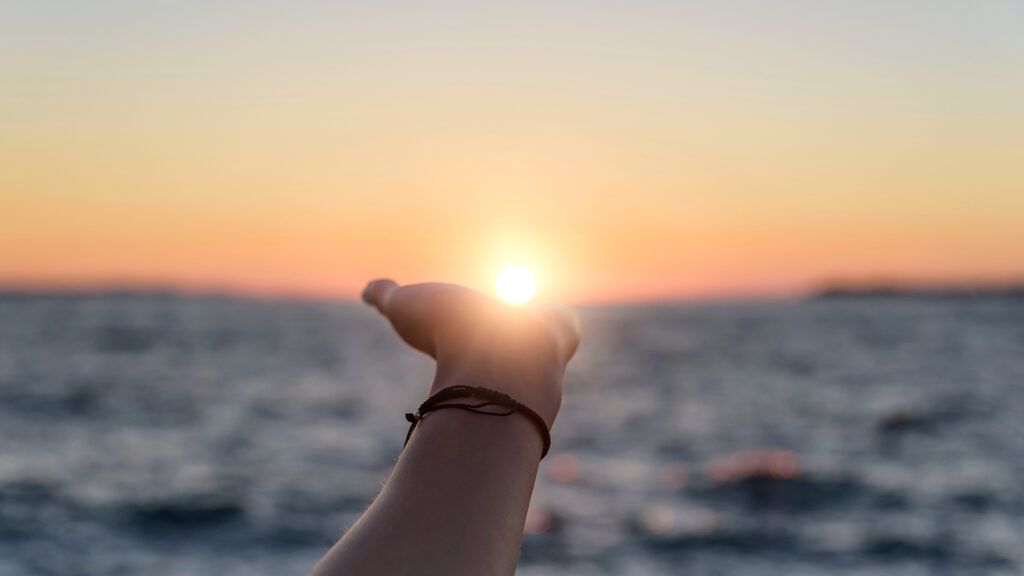 A woman's hand reaches out toward the sun rising over the ocean