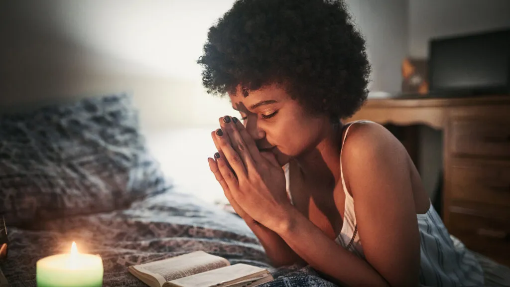 A woman kneeling beside her bed in prayer.