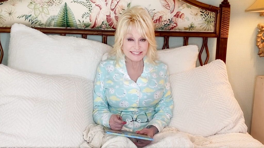 Dolly Parton sits on a bed in PJs, children's book in hand