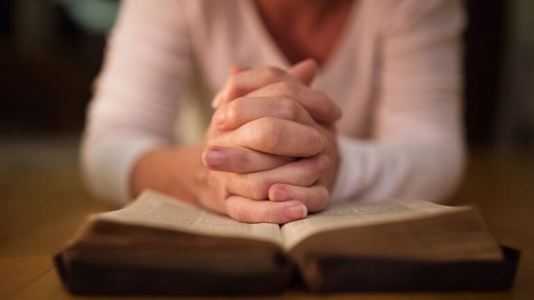 A woman's hands, clasped in prayer, rest on an open Bible