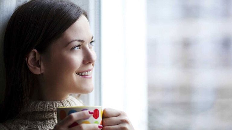 A smiling woman holding a coffee mug gazes out a window