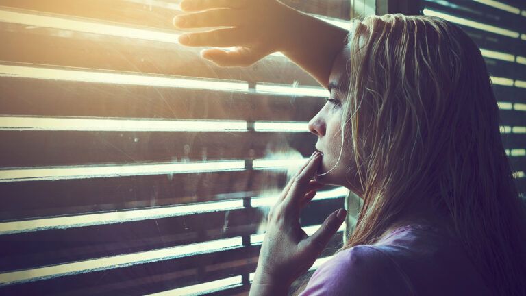 A worried woman peeks through blinds