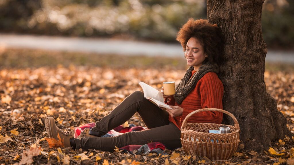 A woman reading outside with fall foliage scenery.