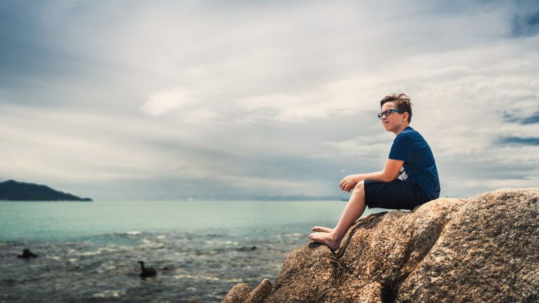 A boy sits on a cliff by a lake