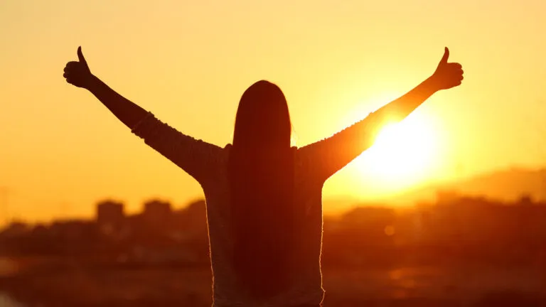 Backlight of a woman raising arms with thumbs up; Getty Images