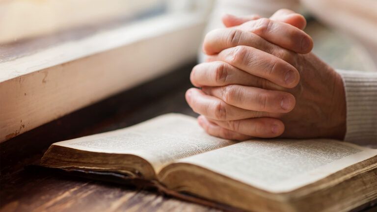 Hands, clasped in prayer, resting upon an open Bible