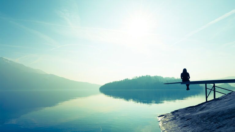 A woman sits quietly by a misty lake at sunrise
