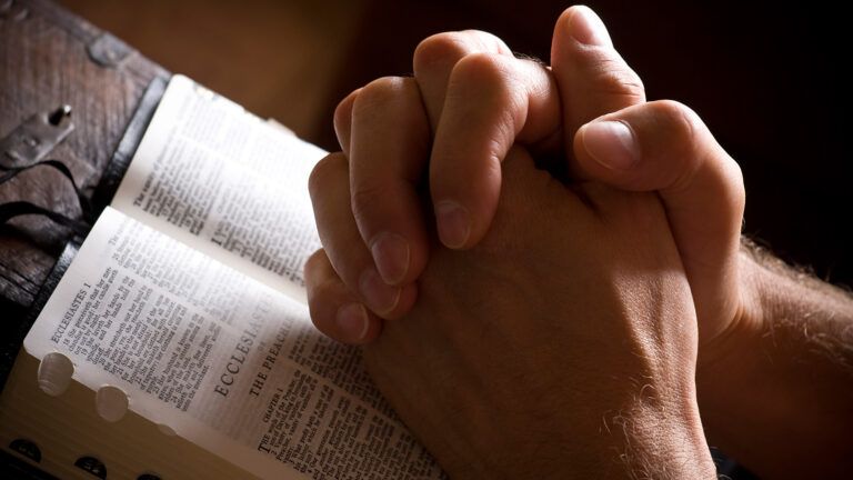 A man's praying hands rest on a Bible