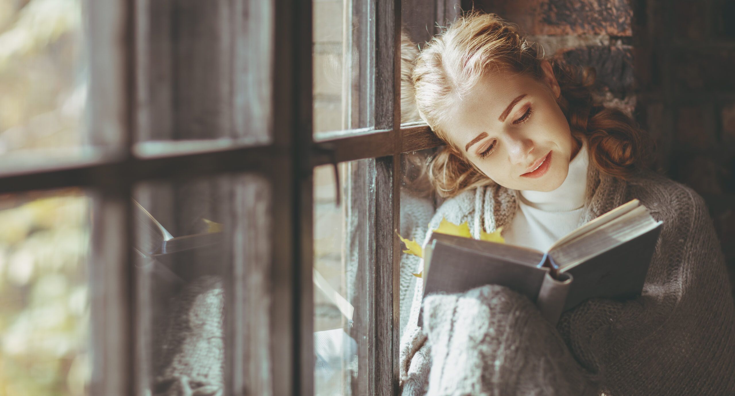 Happy young woman reading book by window in fall (Getty)