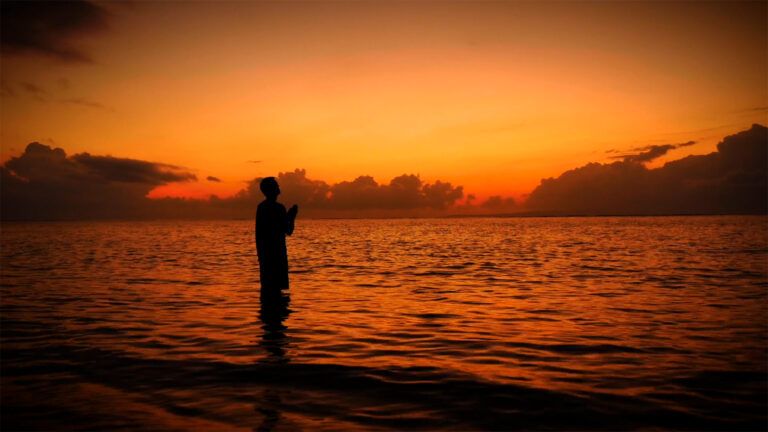 A spiritual pilgrim greets the sunrise while standing in shallow water