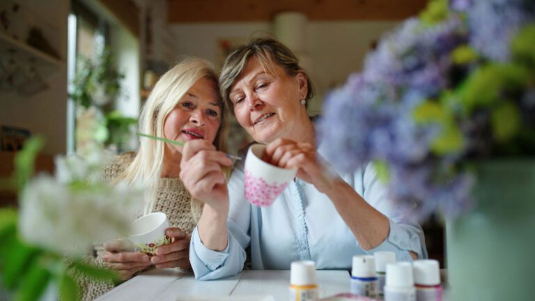 A happy woman artist teaching her friend how to paint a ceramic cup.