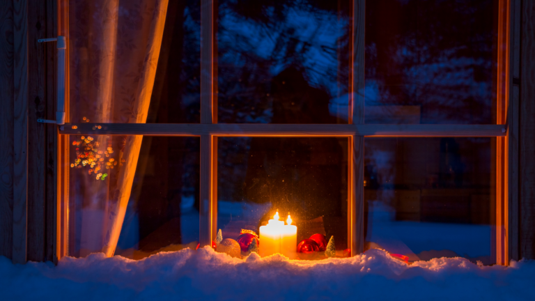 Snowy evening window of a wooden house. On the windowsill are Christmas decorations and burning candles