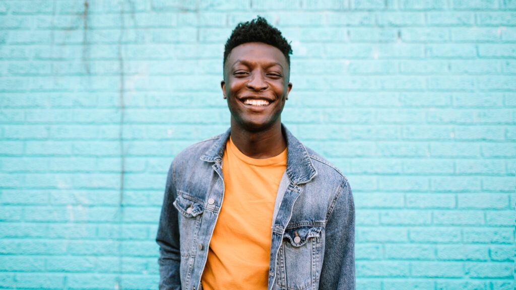 A man in a jean jacket in front of a blue wall smiles because of his positive habits