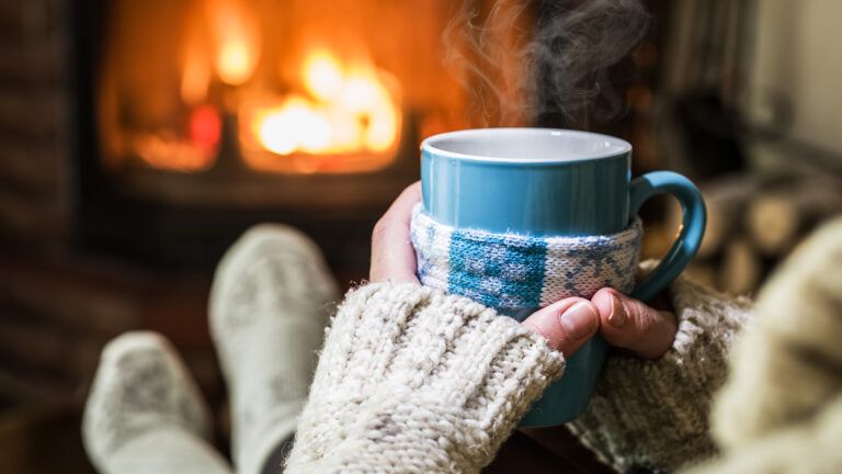 Woman holding a hot cup of coffee by the fireplace; Getty Images