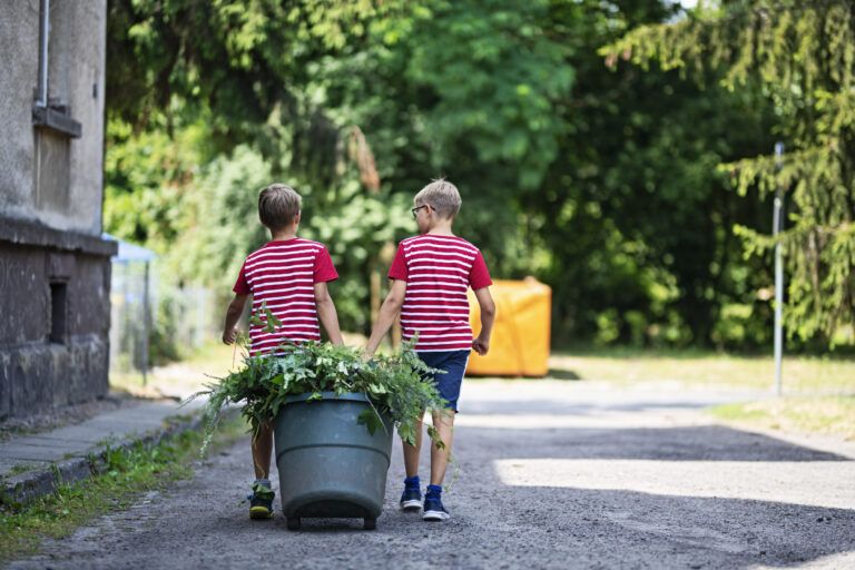 Two boys carrying weeds and pruned branches. 