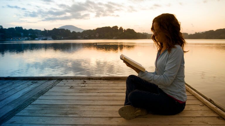 A woman reads the Bible beside a lake at sunrise